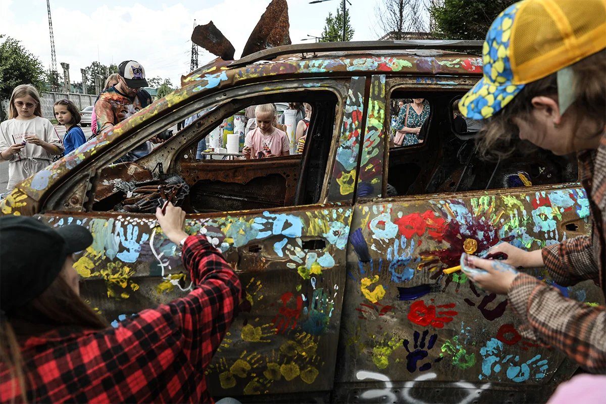 Artists and children paint over a car destroyed in the shelling of Bucha, 30 June 2023. Photo: Oleg Petrasiuk / EPA-EFE