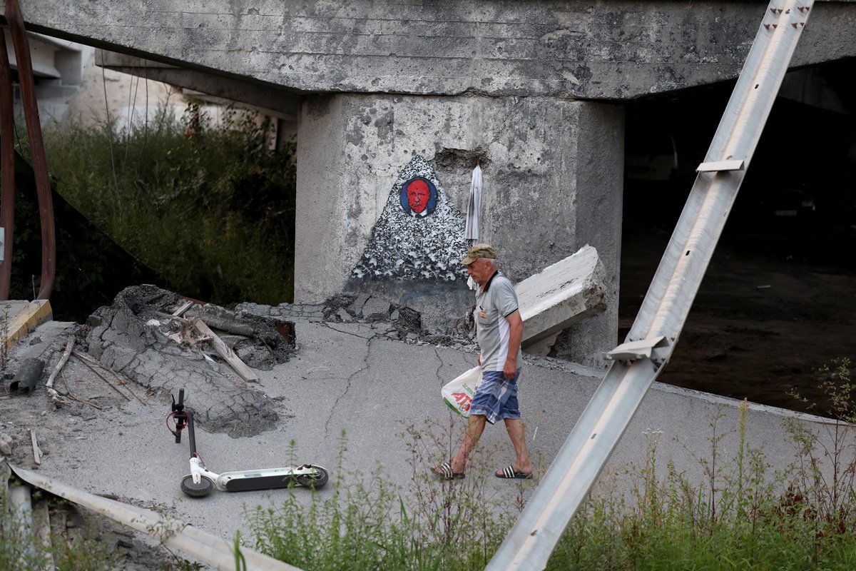 Мужчина идет под разрушенным Ирпенским мостом рядом с фреской, изображающей Владимира Путина, 26 августа 2023 года. Фото: Cathal McNaughton / EPA-EFE